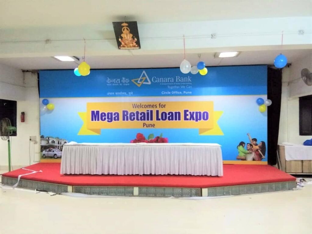 Large metal framed stage backdrop of Canara Bank an elevated platform with red carpet and a long table in the foreground