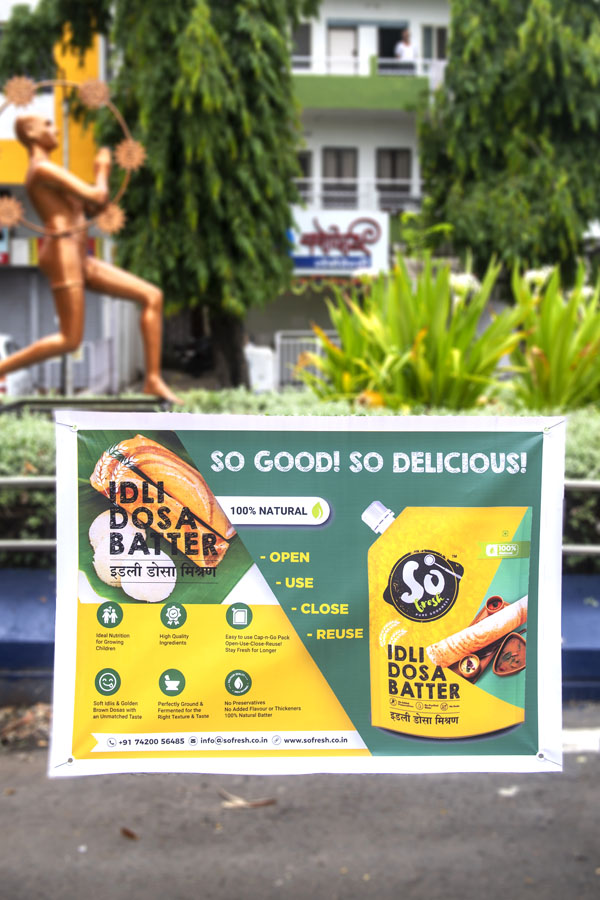 Outdoor banner printed on flex media with eyelets in the corner. This weather proof advertising print is strung up in a garden. Print shows the ad for a dosa batter company