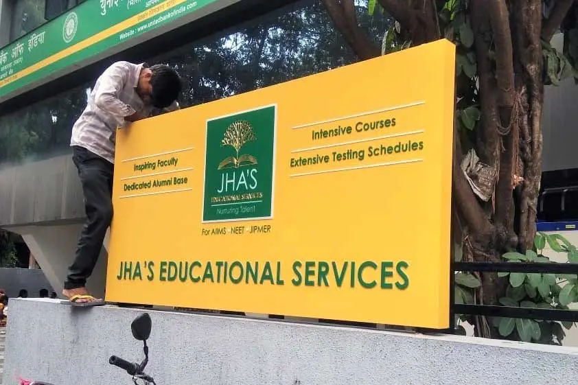 Glow sign board for Jhas Educational Services made of in-cut acrylic letters fixed on an yellow ACP baseboard being fitted on top of a compound wall