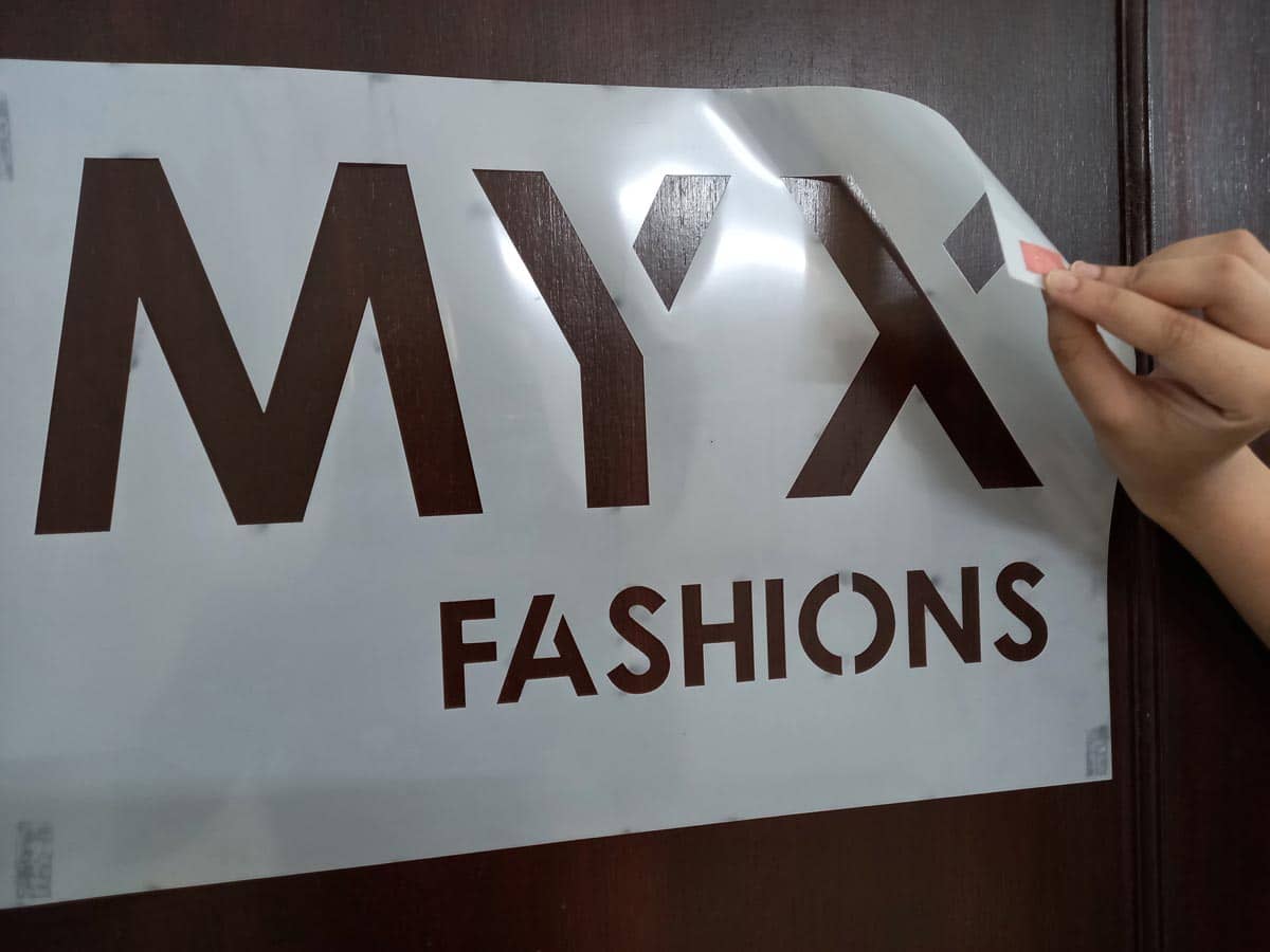 A flexible laser cut custom stencil made of polyester / mylar material is being stuck to a wall using two sided tape. This stencil has the MYX company logo cut out