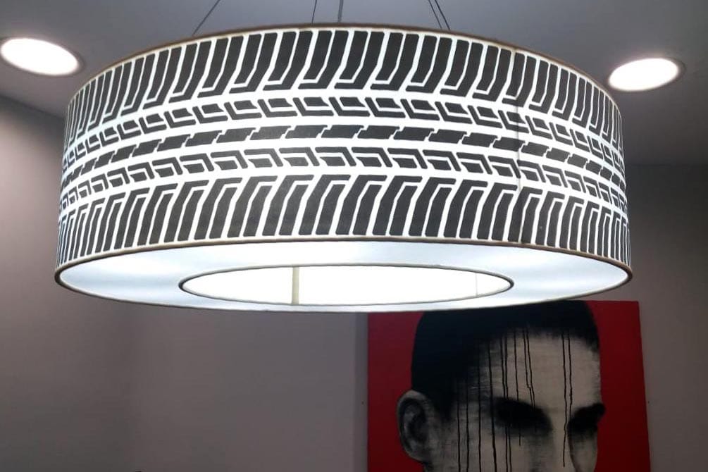 A brightly lit lampshade suspended from the ceiling showing a tyre threads pattern. This design is printed on the fabric stretched on the outside frame of the lamp. There is another framed canvas print on the wall behind it.