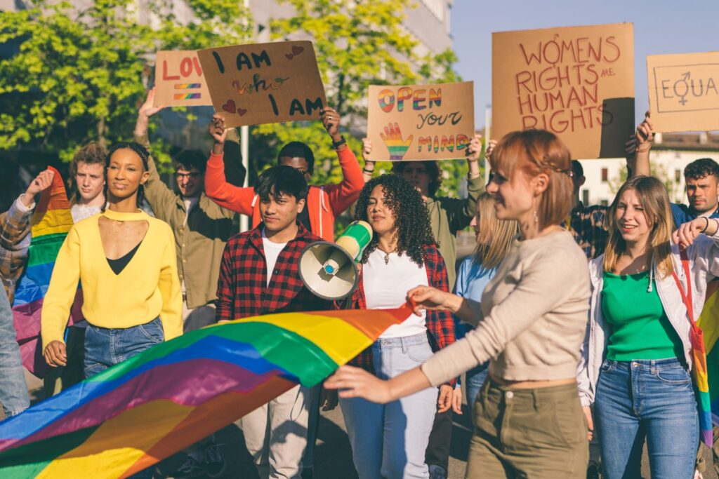 Activists marching for lgbtq+ rights and women's rights in the street holding cardboard placards signs and a rainbow coloured flag