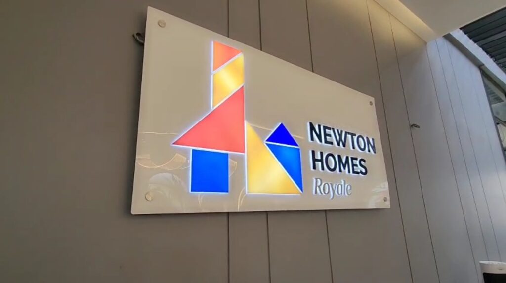 An acrylic logo and company letters are mounted on a white acrylic base plate that is drilled into a wall inside an office. The signboard features embedded LEDs, allowing the logos and letters to shine brightly.