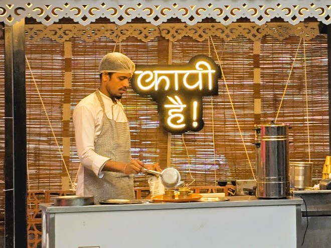 an brightly glowing neon sign made of acrylic hung behind the coffee counter in a restaurant with a barista preparing coffee in the foreground