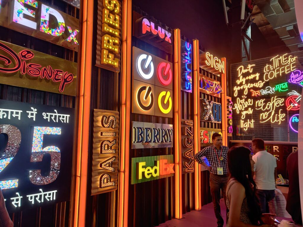 Very colourful glow sign boards made from acrylic, metal, and ACP materials, manufactured in 3D and illuminated with brightly glowing LEDs, All the logo signs are installed next to each other on a wall in a sign shop.