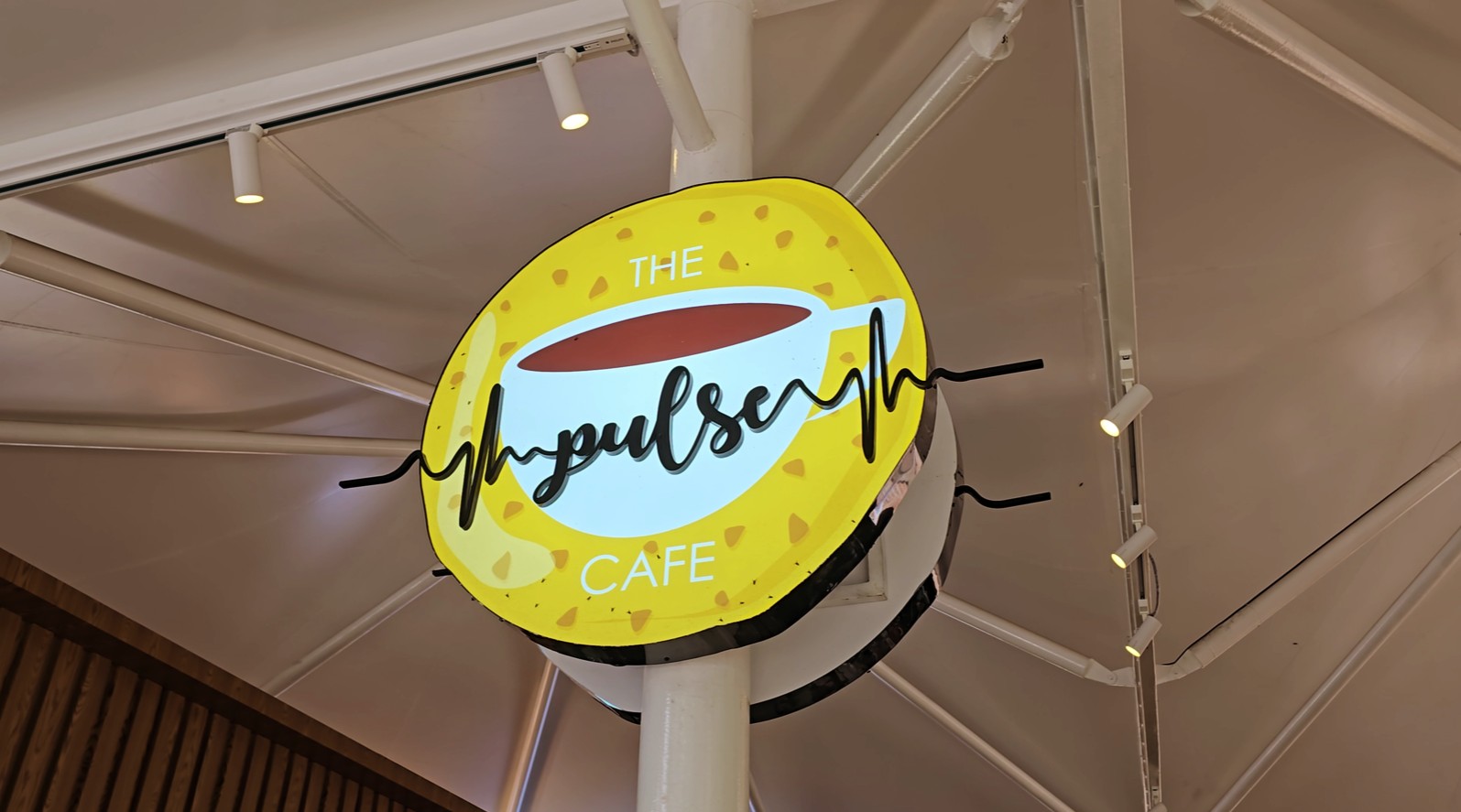 A circular LED acrylic sign board is installed on a round metal pillar inside the cafe. The name of the cafe, "Pulse Cafe," is made from black acrylic and raised in 3D. These acrylic letters are attached to another acrylic sheet, which features an image of a coffee cup printed on it. The entire signboard has embedded LEDs that provide a bright glow.