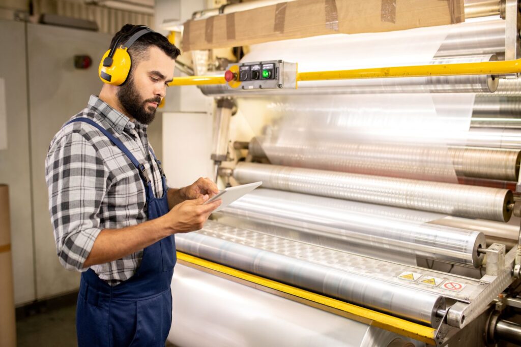 quality control engineer using a tablet to check parameters of processed plastic rolls while standing next to a production machine