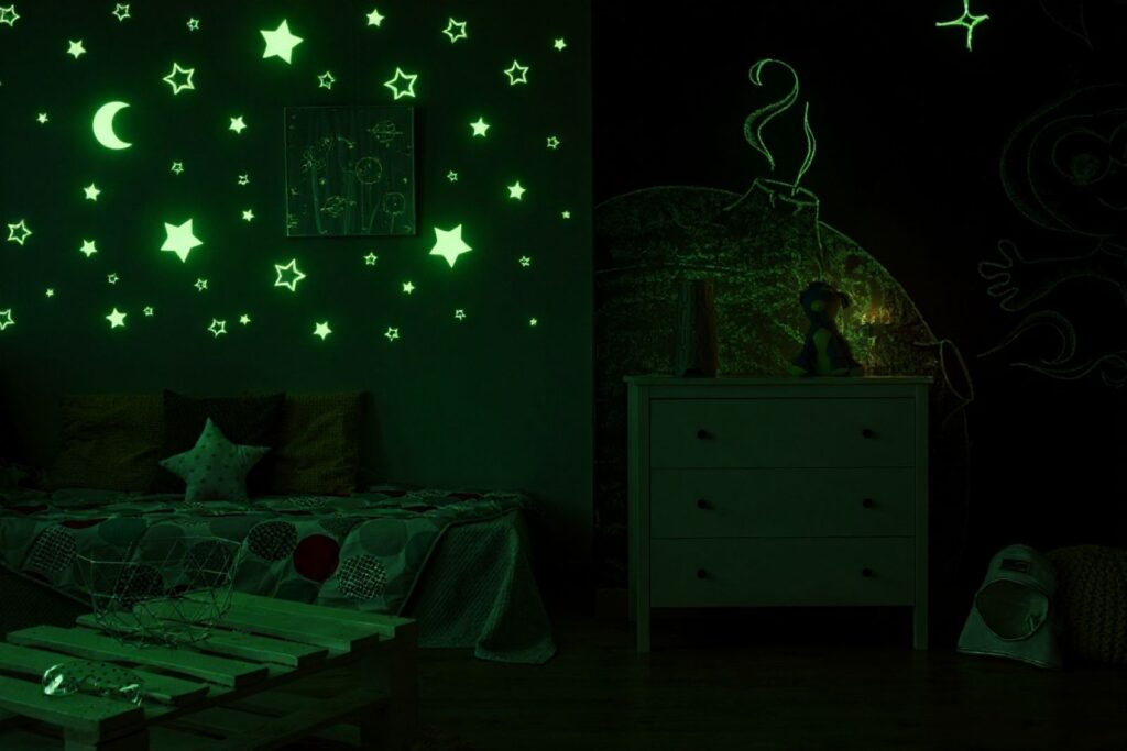 A small child's bedroom featuring stickers of stars and a moon adorning the wall behind the bed, with a sketch drawn on the wall behind the bedside table. The stickers are glowing with a bright greenish light since they are made out of glow-in-the-dark material. This image has been touched up in Google Gemini using AI.