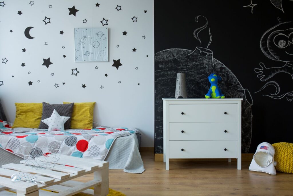 A small child's bedroom featuring stickers of stars and a moon adorning the wall behind the bed, with a sketch drawn on the wall behind the bedside table.