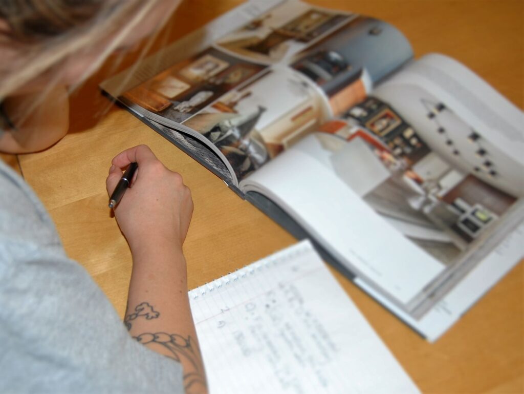 Over the shoulder view of a person viewing a brochure opened out on a wooden table The catalog has a hard case binding cover and the person is taking notes on a notepad