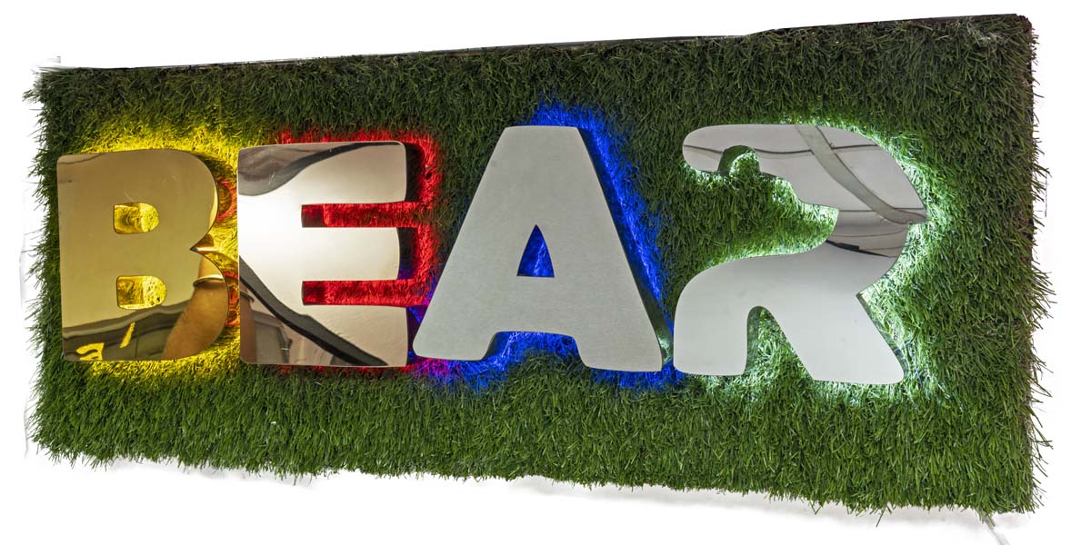 Metal letter sign board with back glow lights mounted on a grassy surface. Each letter having different finishes like gold, rose gold, chrome and brush finish steel