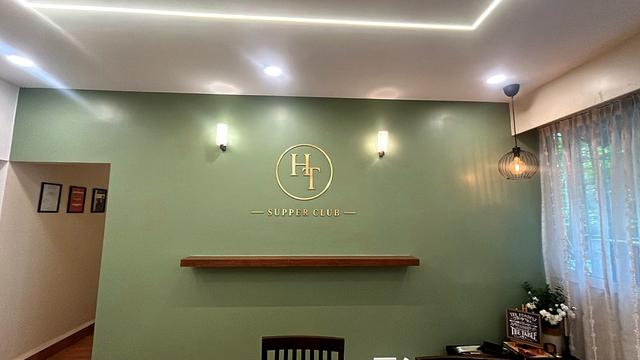 Wide view of the Her Table Supper Club cooking studio in Pune: a sage-green feature wall with a small gold acrylic monogram, wall sconces, a wooden shelf, a pendant lamp and sheer curtains.