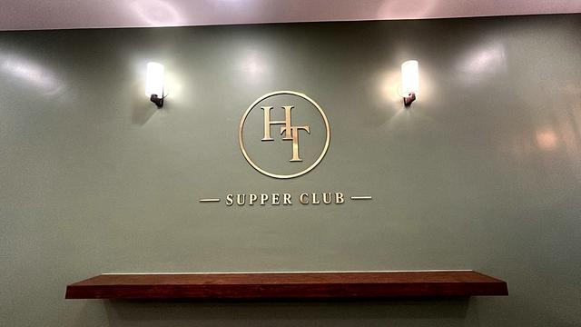 Gold acrylic HT monogram in a circle above the words Supper Club, fixed directly onto a sage-green painted wall with no base plate, between two wall sconces and above a wooden shelf, at Her Table Supper Club cooking studio in Pune.