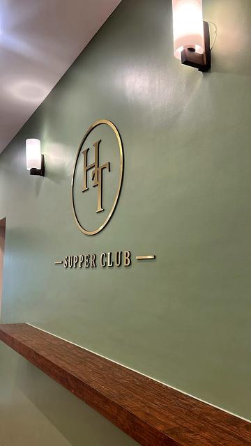 Portrait view looking up at the gold acrylic HT monogram and Supper Club lettering on a sage-green wall, lit by warm wall sconces, with a wooden shelf in the foreground.