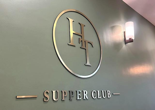 Close-up at an angle of the gold acrylic HT monogram and thin circular ring on a sage-green wall, showing the thickness of the layered letters and the soft shadow they cast, with a wall sconce beside it.