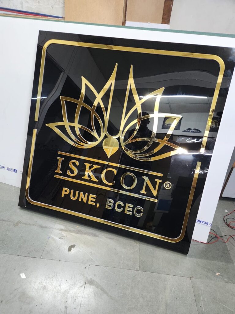 Gold mirror-finish laser-cut acrylic ISKCON Pune BCEC logo board on a black acrylic base, daylight shot
