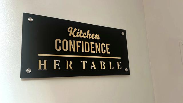 Matte black acrylic nameplate reading Kitchen Confidence and Her Table in raised gold acrylic letters, fixed to a white wall with four chrome corner caps.