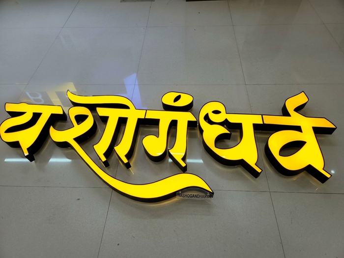 Illuminated orange acrylic-faced aluminium channel letters spelling Yashogandharva, laid out and lit up in the Orchid Digitals workshop for testing before the ceiling-suspended installation