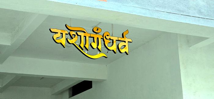 Daytime view from below showing the Yashogandharva channel letters hanging from thin metal rods fixed to the ceiling, with no visible support frame or wall