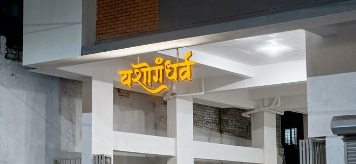 Close-up night shot of the orange-lit Yashogandharva channel letters glowing under the building's entrance canopy, appearing to float with no visible support