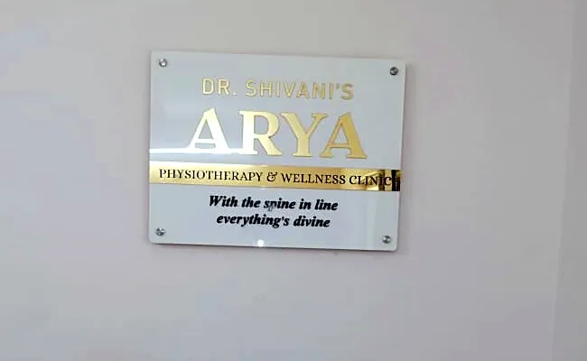 ARYA Physiotherapy & Wellness Clinic name board on the clinic wall: gold acrylic letters on a white acrylic base plate with stand-off studs