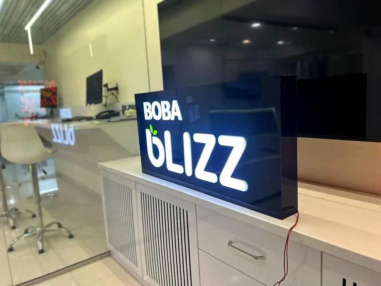 The Boba Blizz sign lit up: white 3D acrylic letters and green leaves glowing on a glossy black acrylic box, made in Pune for a client in the USA