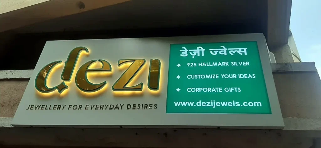 Dezi Jewels shop board: gold 3D letters with a warm LED back glow on white ACP, a green panel with the name in Marathi