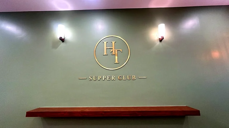 Brushed gold acrylic HT Supper Club monogram fixed directly on a sage green wall, between two wall lamps