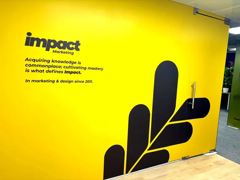 Impact Marketing office in Pune: glass cabin partitions and door covered in a yellow brand wall on laminated opaque film, with the Impact logo, their line and a dark leaf mark