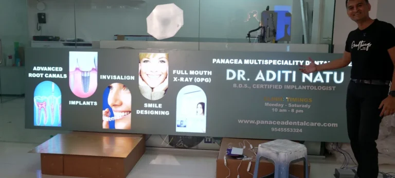 The 10 ft lit sign board for Dr Aditi Natu’s Panacea Multispeciality Dental Care switched on: five glowing photo windows and the clinic name on a silver-grey ACP base