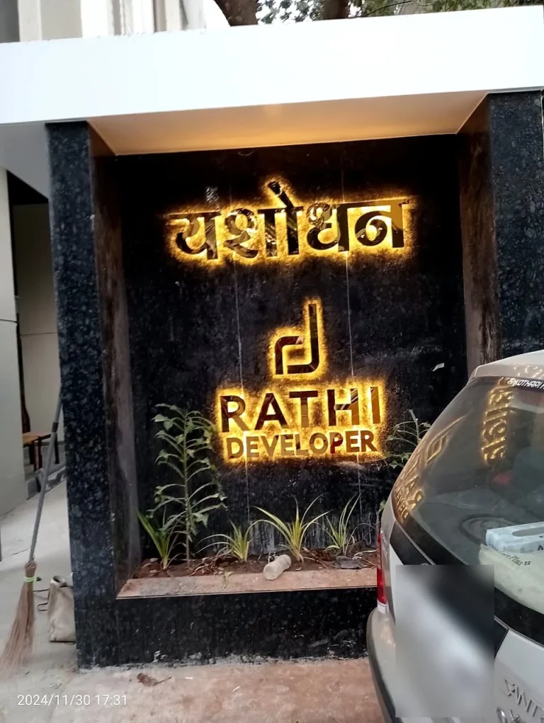 Yashodhan in Marathi and the Rathi Developer logo in gold steel letters glowing warm white on the granite gate wall at night, Aundh, Pune