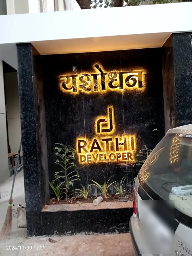 Yashodhan in Marathi and the Rathi Developer logo in gold steel letters glowing warm white on the granite gate wall at night, Aundh, Pune