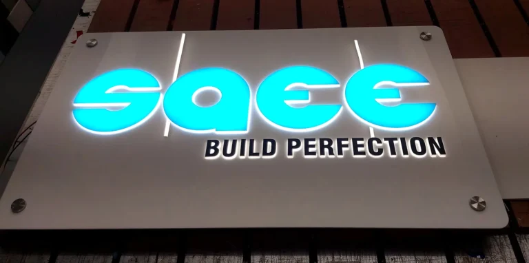 SAEE BUILD PERFECTION backlit acrylic logo board lit in our workshop: blue acrylic letters glowing on a white acrylic base plate with steel stud caps at the corners
