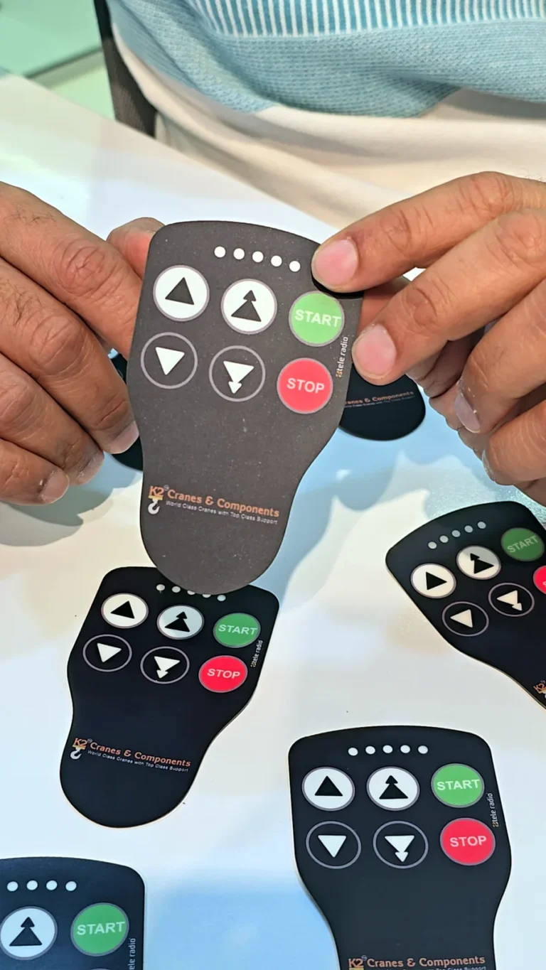 Printed control panel overlay for a Tele Radio crane remote, branded K2 Cranes & Components, held up over a table of finished overlays